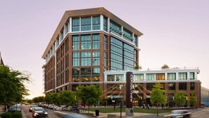 A modern building with a glass facade, a prominent entrance with a sign, and a parking lot with cars.