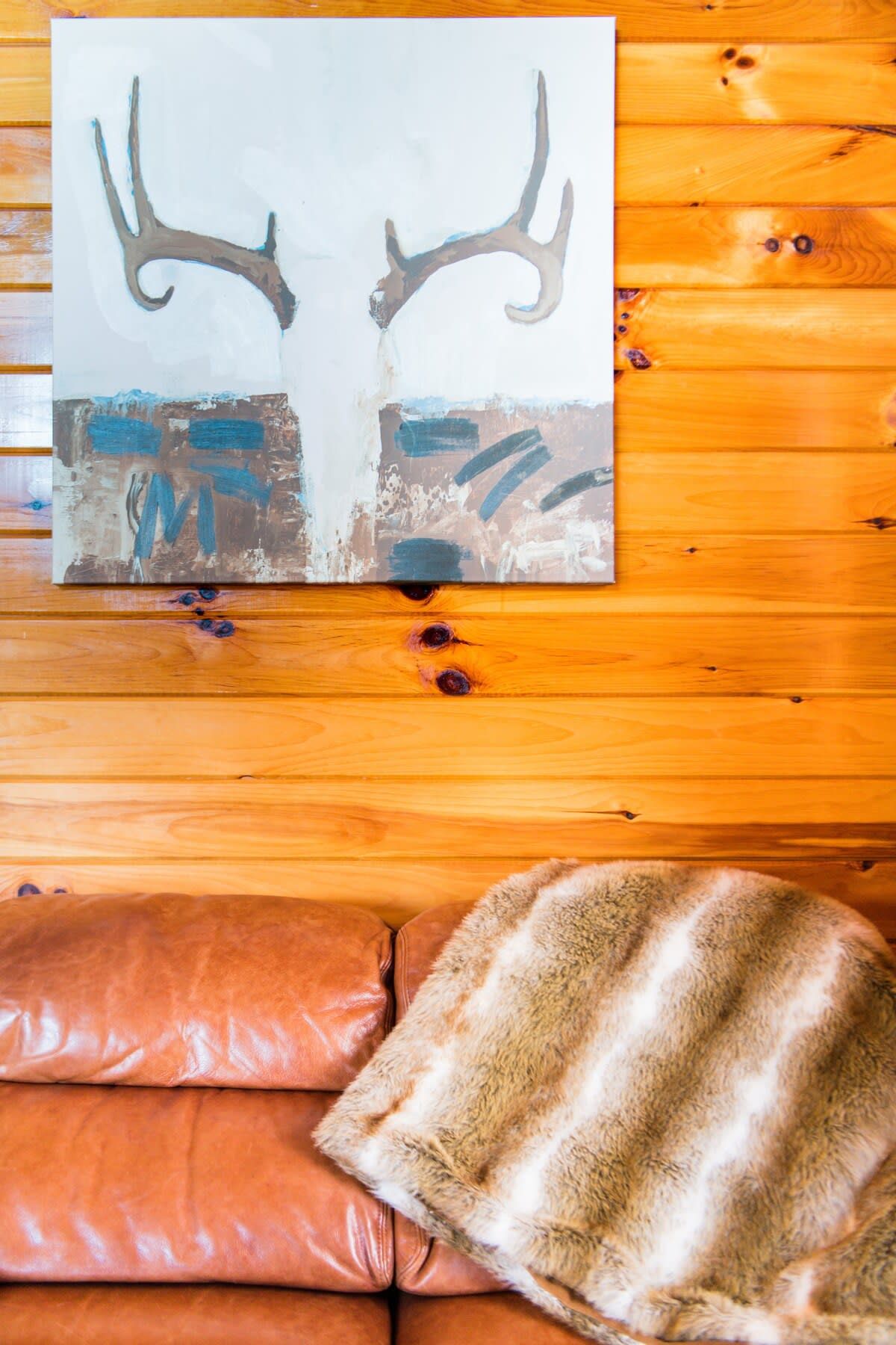 Interior de una cabaña de madera con un cuadro de cuernos de ciervo en la pared, un sofá de cuero y un plaid de piel.