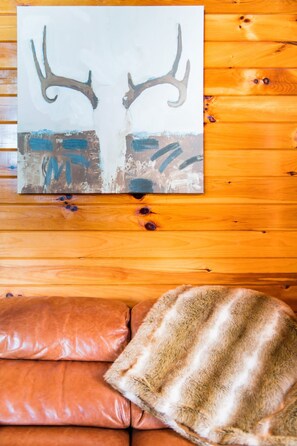 Interior de una cabaña de madera con un cuadro de cuernos de ciervo en la pared, un sofá de cuero y un plaid de piel.