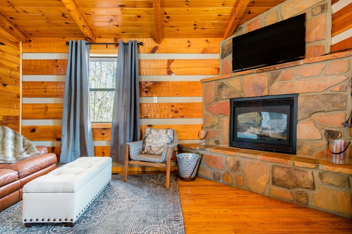 Ein gemütliches Blockhauss-Interieur mit Kamin aus Stein, Holzwänden, einem Ledersofa und einem an der Wand befestigten Fernseher.