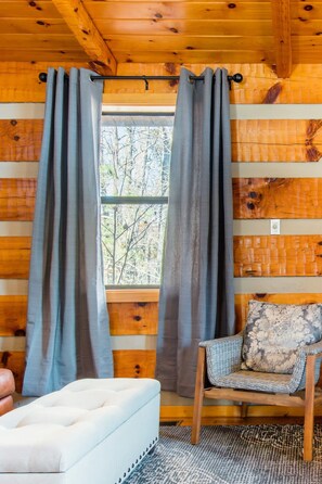 Interior d'una cabana amb finestra, cortines, una cadira i un sofà.