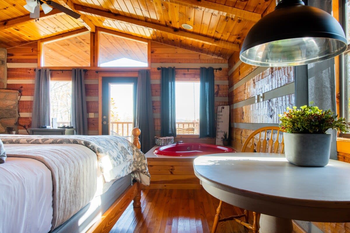 A cozy cabin-style room with a bed, a round table, a potted plant, and a view of the outdoors through large windows.