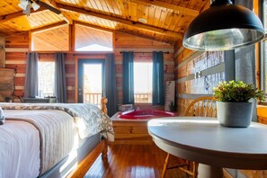A cozy cabin-style room with a bed, a round table, a potted plant, and a view of the outdoors through large windows.