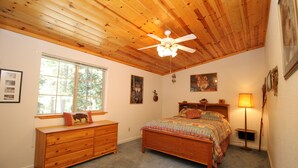 Una camera da letto con soffitto in legno, un letto con copriletto colorato, un comò, una finestra e una lampada fissata al muro.