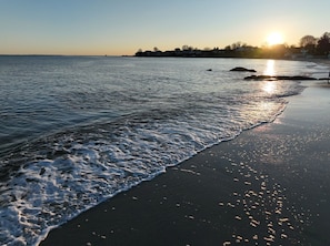 A beach with waves gently lapping onto the shore, a clear sky, and the sun setting in the distance.