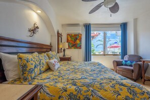Hotelkamer met een bed, een stoel, een raam met gordijnen, een plafondventilator en een luchtverkoeler aan de muur.