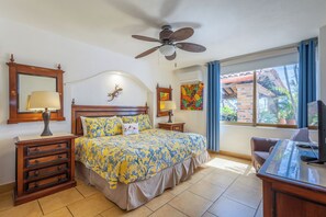 Un dormitorio con una cama, un escritorio, una silla, un ventilador de techo, una ventana con cortinas y un espejo montado en la pared.
