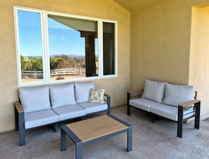 A balcony with two sofas and a wooden table, large windows with a view of a fenced area and hills.