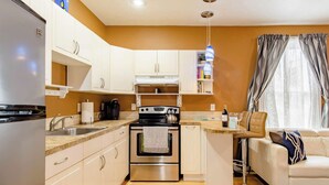 A compact kitchen with a stove, oven, and sink, a small island with a wine bottle, and a sofa in a living area with a window. - 1880 Victorian In Heart Of Downtown  (St. Augustine)