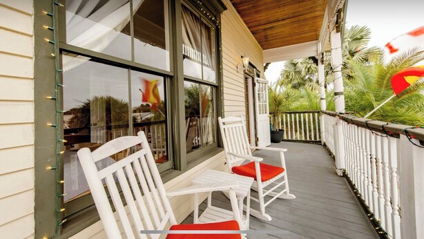 A covered porch with white chairs and a table, a wooden ceiling, and a view of palm trees and a flag. - 1880 Victorian In Heart Of Downtown  (St. Augustine)