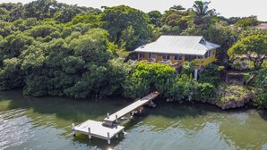 Een huis omringd door weelderige begroeiing, een steiger die in het water uitloopt, en een heldere lucht.
