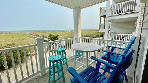 Katettu ulkotila, jossa on pöytä ja tuoleja, ja josta avautuu näkymä rannalle ja dyynimaisemaan.