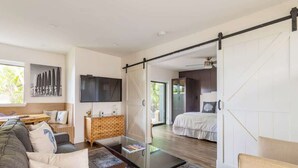 Ein modernes Wohnzimmer mit einem Glastisch, einer Couch und einem Fernseher. Eine Schiebetür führt zu einem Schlafzimmer mit einem Bett und einem Badezimmer.