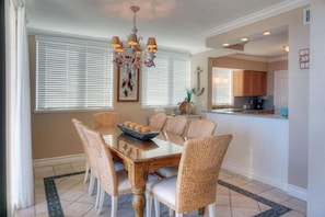 A dining area with a wooden table and wicker chairs, a chandelier, and a kitchen in the background.