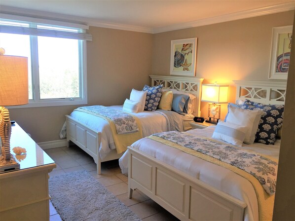 A bedroom with two beds, a nightstand with a lamp, a window with blinds, and a painting on the wall.
