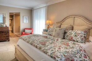 A bedroom with a large bed, a wooden dresser, a red chair, and a nightstand with a lamp.