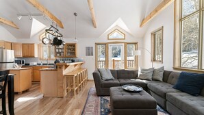 Ein großzügiger Wohnbereich mit einem großen grauen Sofa und Hocker, Holzboden und einer Küche mit Holzschränken und einer abgehängten Küchenleiste.