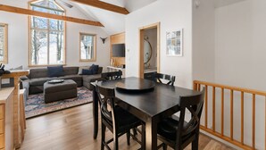 Ein modernes Wohnzimmer mit einem dunklen Esstisch aus Holz, einem grauen Sofa und einem großen Fenster mit Blick auf Bäume.
