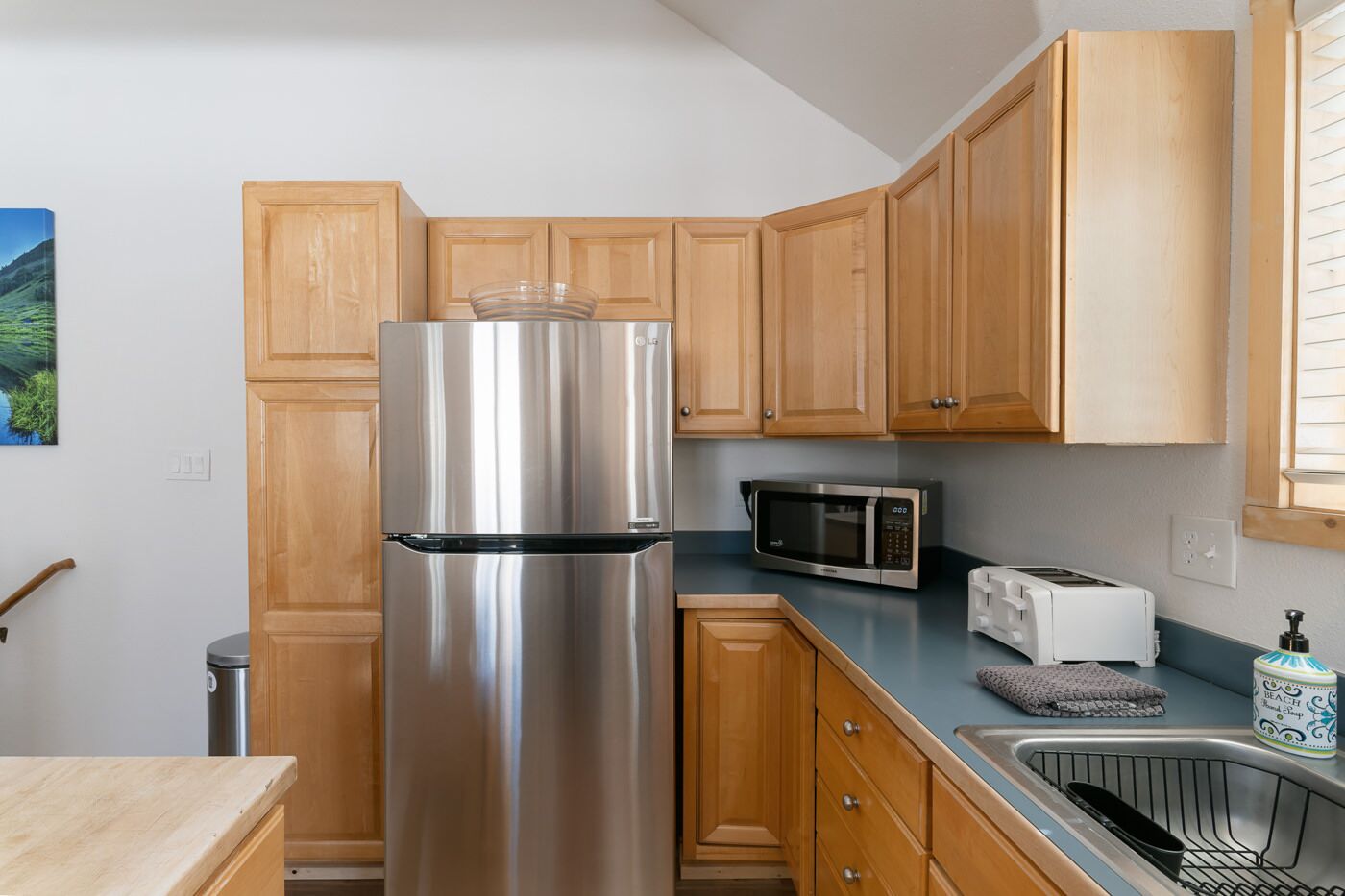 A compact kitchen with wooden cabinets, a stainless steel refrigerator, a microwave, a toaster, and a sink.
