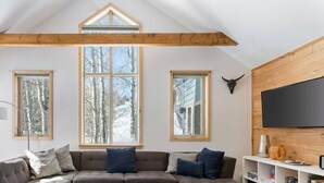Ein modernes Wohnzimmer mit einem Ecksofa, großen Fenstern, einem wandmontierten Fernseher und einem Holzbalken an der Decke.