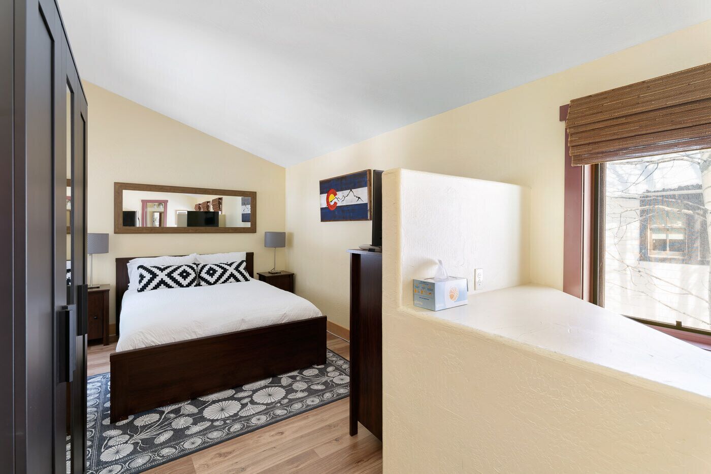 A bedroom with a bed, a bedside table, a lamp, a mirror, a window with a view, and a bedside box of tissues.