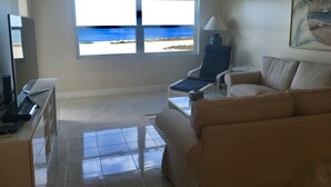 A living room with a view of the beach, a chair, a sofa, a lamp, and a TV.