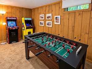 A room with a foosball table, two arcade games, and framed pictures on the wall.
