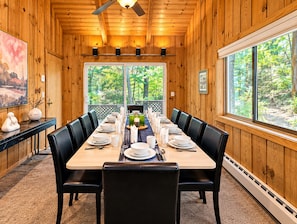 A dining area with a long table set for six, wooden walls, and large windows offering a view of trees.