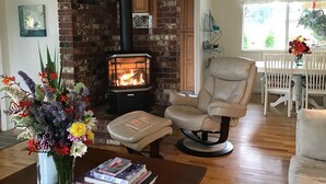 Een woonkamer met een open haard van baksteen, een leren fauteuil, een eethoek met een witte tafel en stoelen, en een bloemenvaas op een tafel.