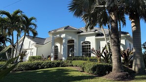 A white house with a grey roof, surrounded by palm trees and well-maintained lawns. - Waterfront Pool Home with Gulf Access (Cape Coral)