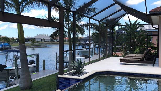 A covered outdoor swimming pool with a view of the water, palm trees, and a dock with boats. - Waterfront Pool Home with Gulf Access (Cape Coral)