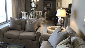 A living room with a grey sofa, a glass coffee table, a chandelier, and a dining area with chairs and a table.