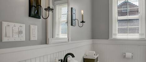 A bathroom with a white sink, vanity, and toilet. There is a mirror above the sink, and a window with blinds on the wall.