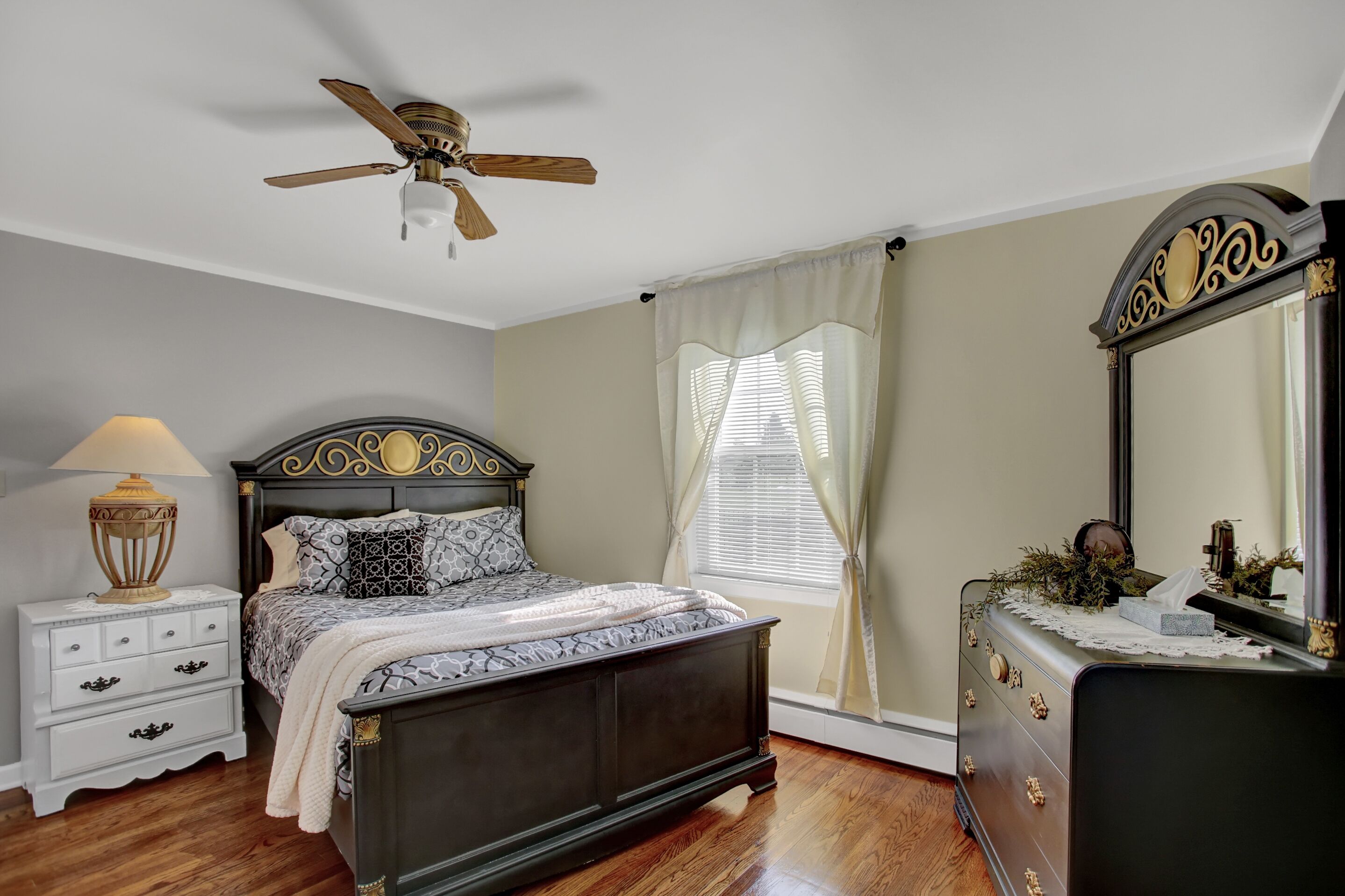 A bedroom with a bed, a nightstand, a dresser, a ceiling fan, and a window with curtains.