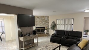 A living room with a black leather sofa, a coffee table, a flat-screen TV mounted on the wall, a fireplace, and a potted plant.