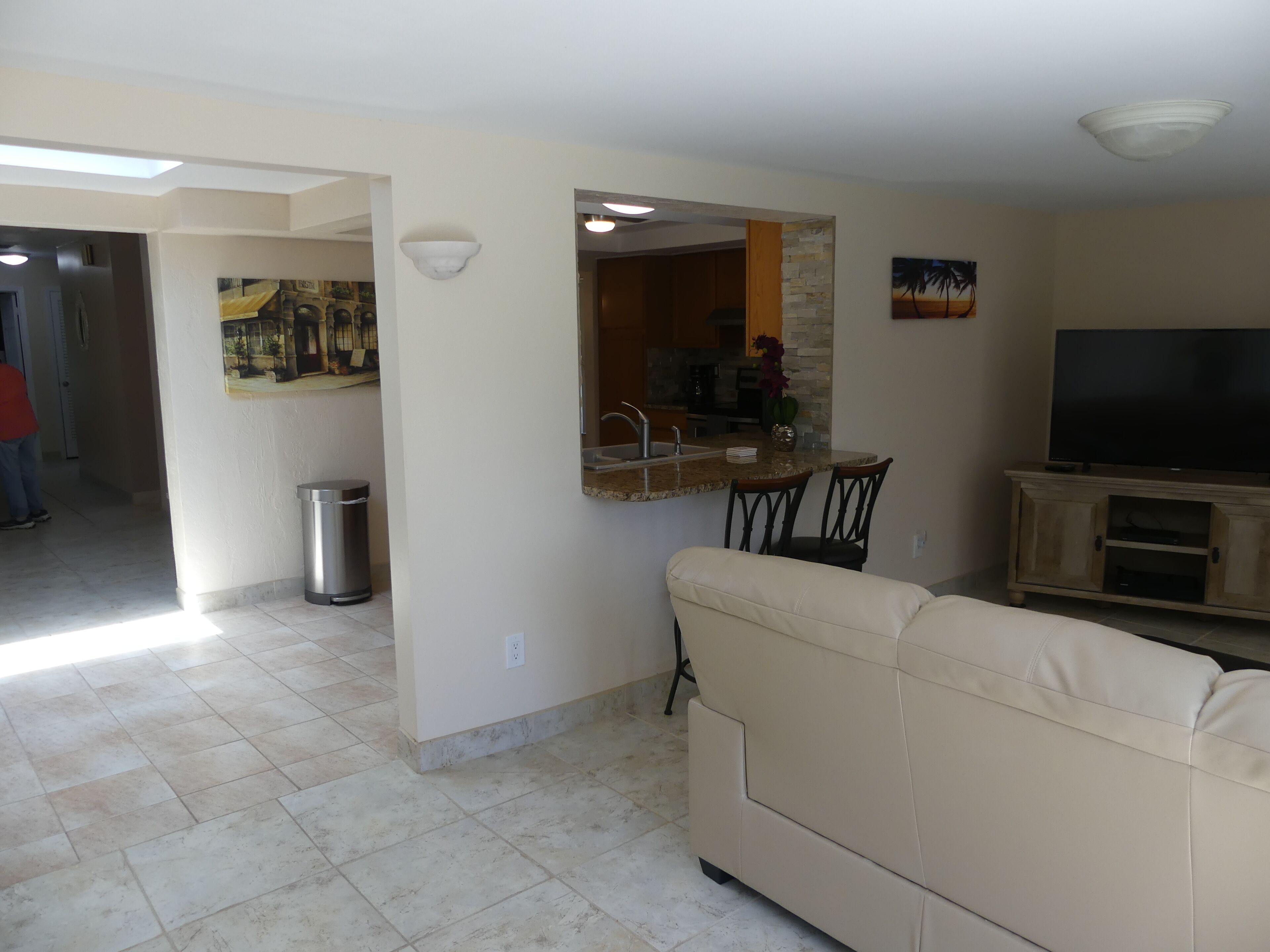 A living room with a beige sofa, a TV, a kitchen area with a stone backsplash, and a trash can.