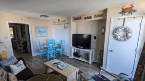 A living room with a TV, a dining table, and blue chairs. There's a door with a wreath and a sign above it. - Beach Lover's Condo With Spectacular Ocean View, Pool, Lazy River, & Jacuzzi!  (North Myrtle Beach)