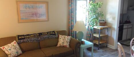 A living room with a brown sofa, a small table with a lamp, a chair, and a framed picture on the wall.
