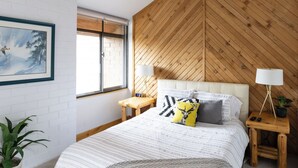 A bedroom with a bed, a window, a wooden wall, a nightstand, a lamp, and a plant.