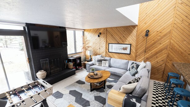 A modern living room with a sofa, coffee table, and foosball table. There is a fireplace, a large window, and a herringbone-patterned wall.