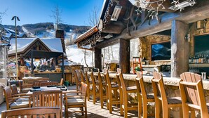 An outdoor seating area with wooden chairs and tables, a bar with bottles, and a view of a snowy mountain landscape. - Luxury Ritz Carlton 2BR Residence - Ski-in/out & Private Laundry (Beaver Creek)