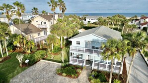 A two-story house with a balcony, surrounded by palm trees and a paved driveway. - Ocean Song – Large 2 Bedroom with Private Ocean View Balcony & Sea Breezes & Pool Access (St. Augustine)