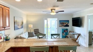 A kitchen with a central island, wooden cabinets, and a ceiling fan. There is a dining area with chairs and a table, and a TV mounted on the wall. - Ocean Song – Large 2 Bedroom with Private Ocean View Balcony & Sea Breezes & Pool Access (St. Augustine)