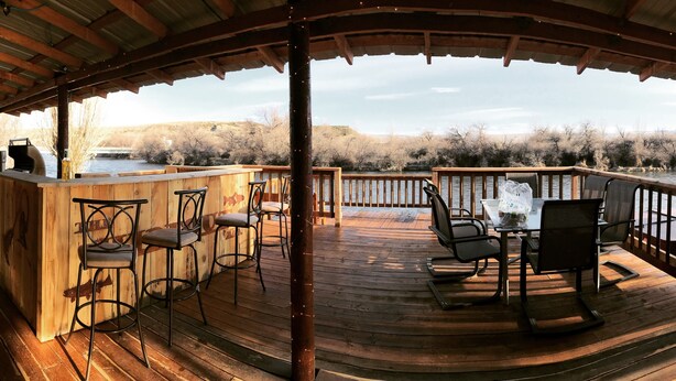 Una terraza de madera con mobiliario exterior, una parrilla y vistas a un río y árboles.