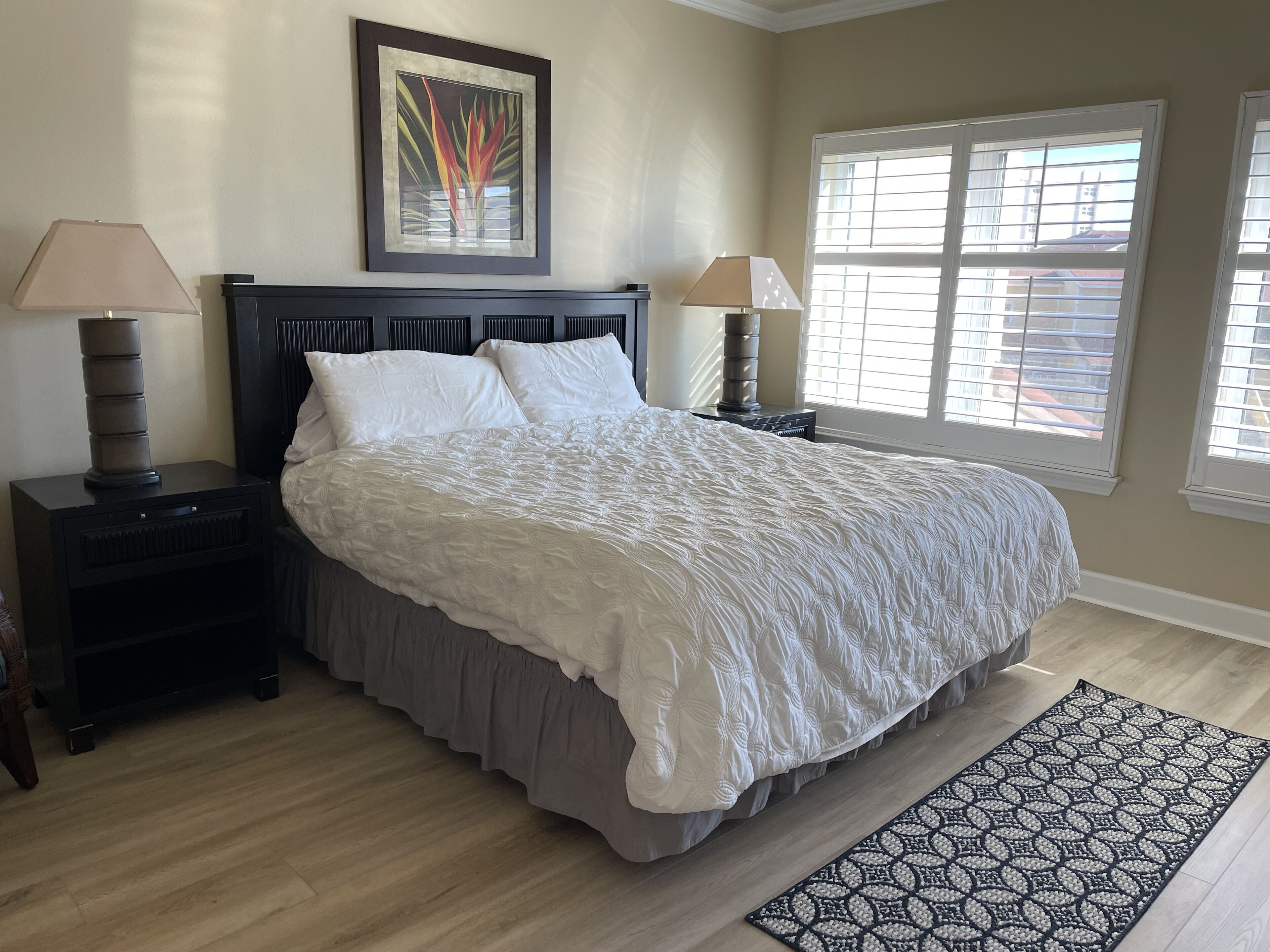 A bedroom with a large bed, two bedside tables, a nightstand, a window with blinds, and a framed picture on the wall.