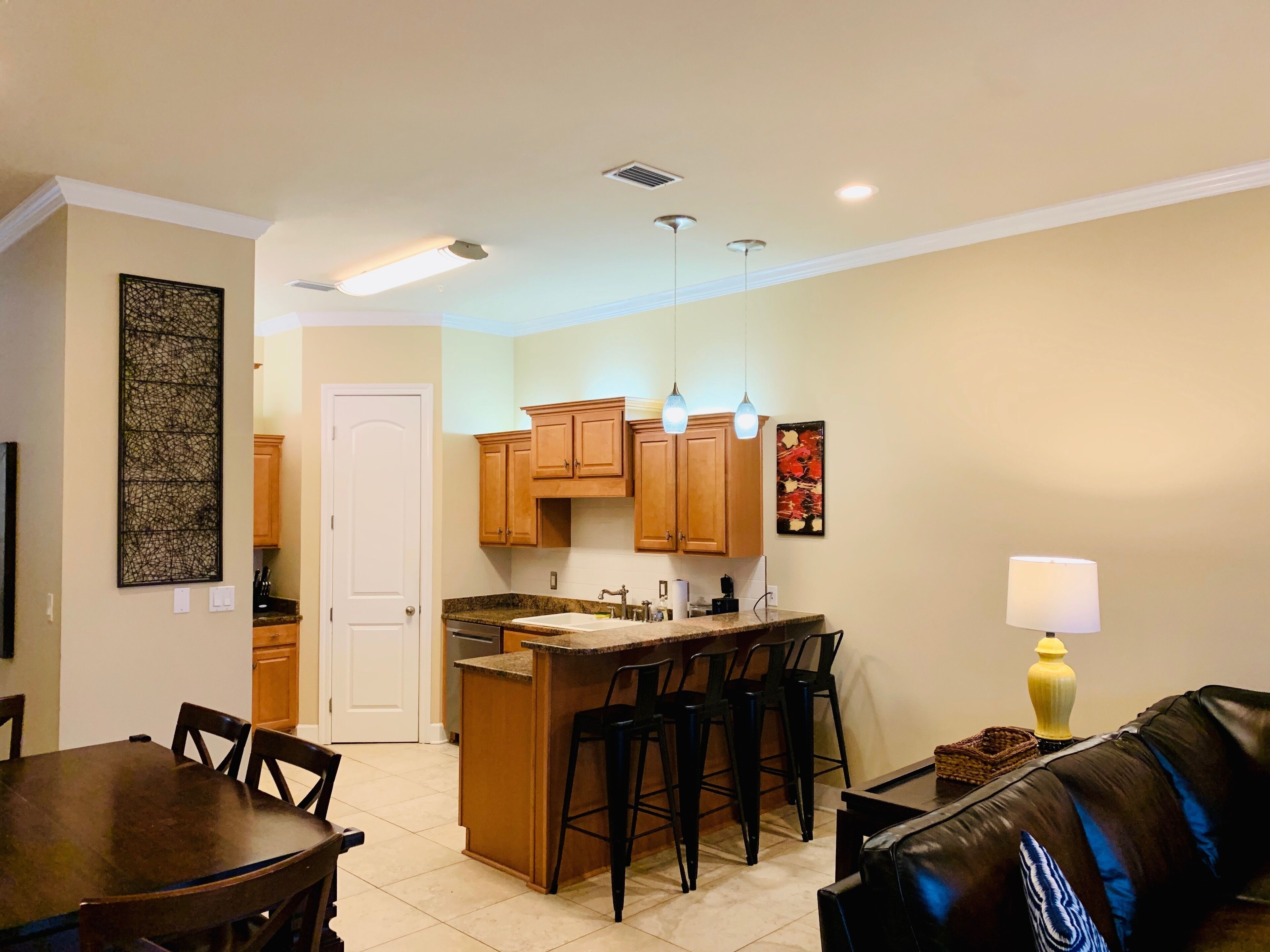 A compact kitchen with wooden cabinets, a central island with bar stools, a dining table with chairs, and a living area with a sofa and a lamp.