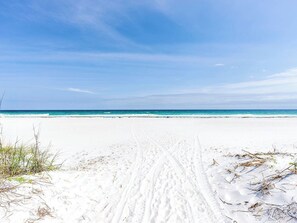 En strand med klart himmel, en sandsti der fører ned til vandet, og sparsom vegetation.