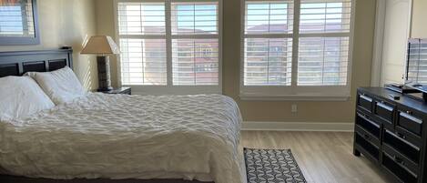 A bedroom with a large bed, two windows with blinds, a dresser, and a nightstand with a lamp.