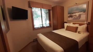 Un dormitorio con cama, una ventana con persianas, un televisor montado y un cuadro en la pared.