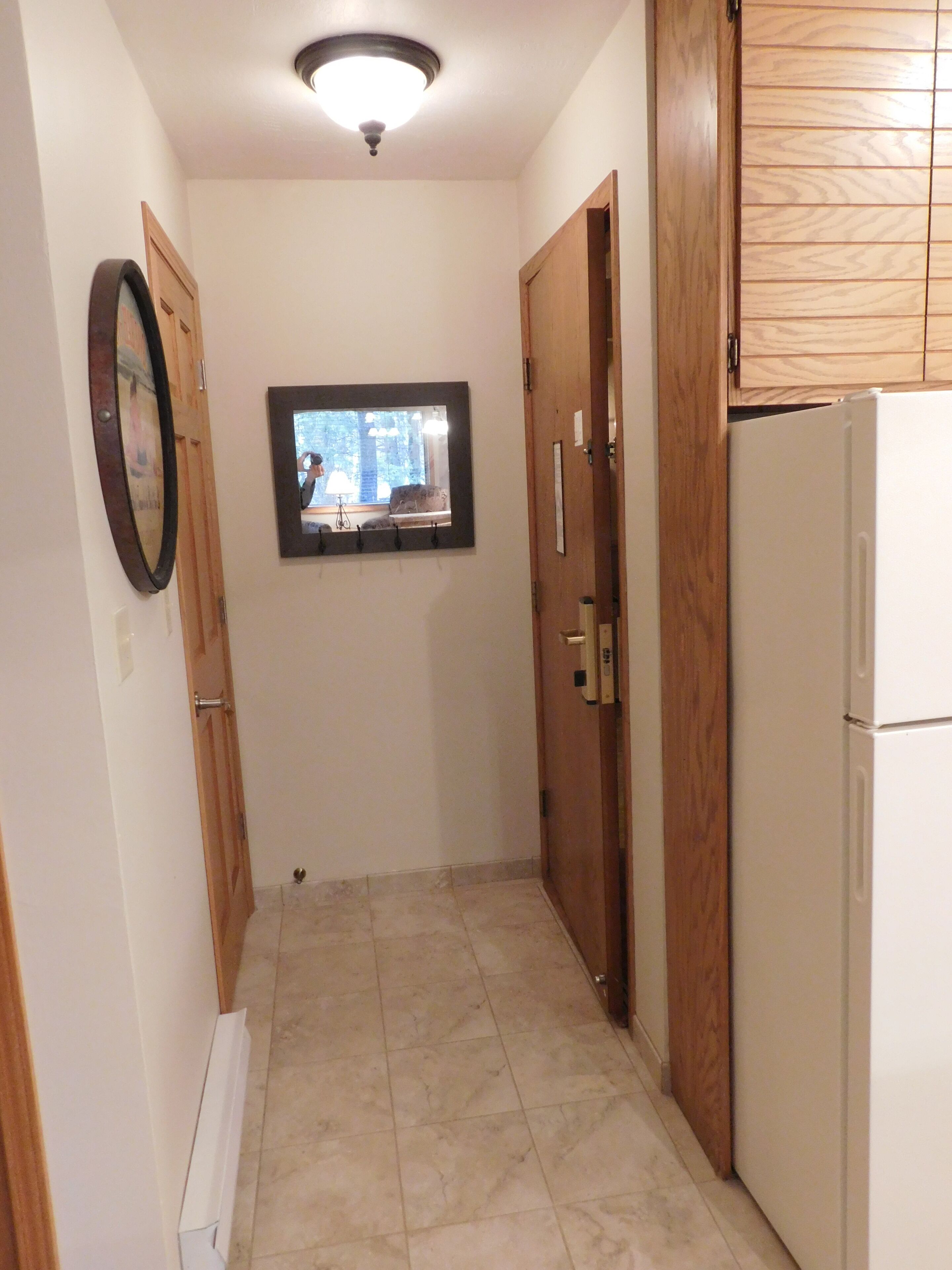 A narrow hallway with a tiled floor, wooden doors, a white refrigerator, and a framed picture on the wall.